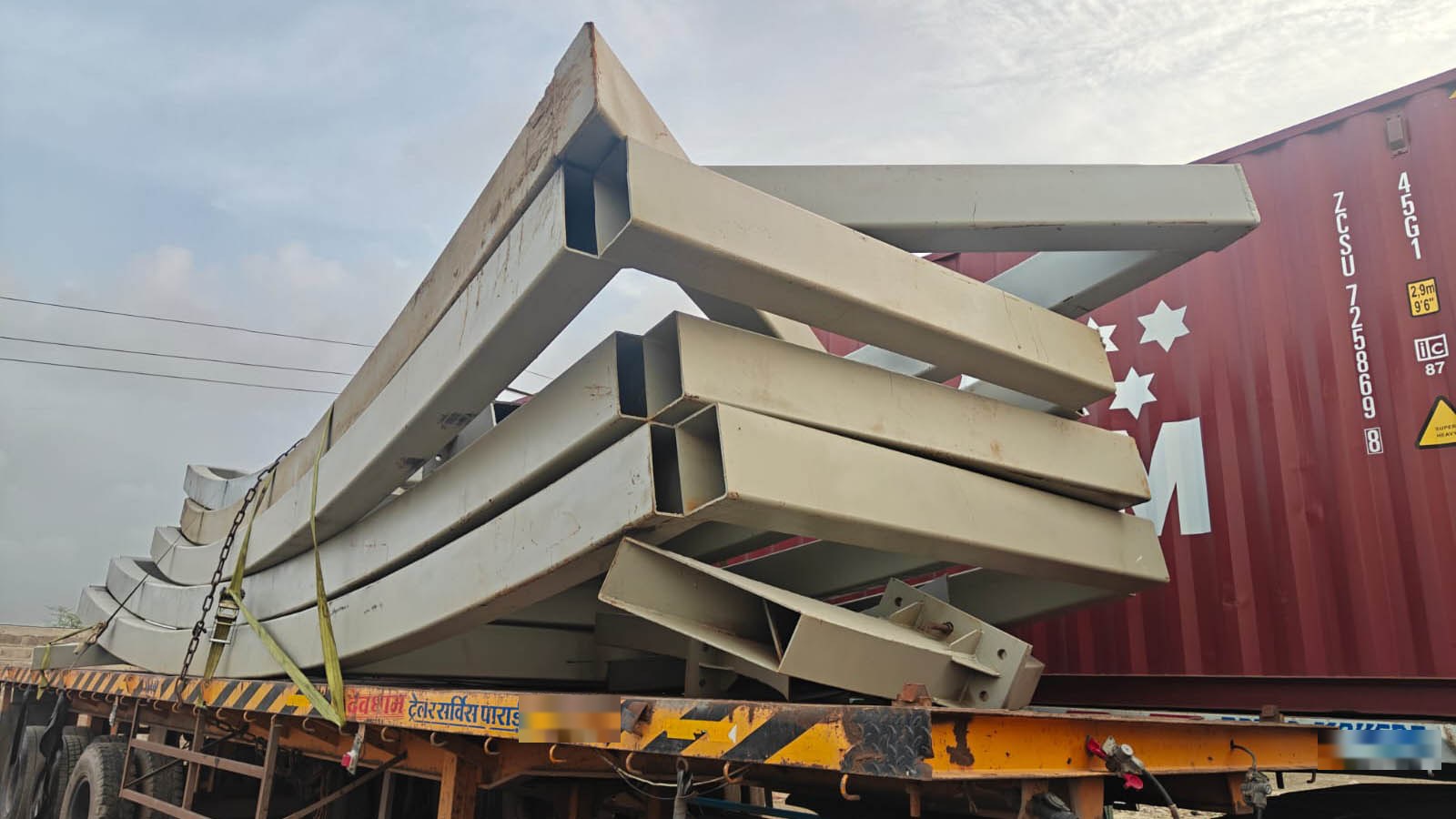 Fabricated steel sections strapped to a flat-bed trailer beside a shipping container